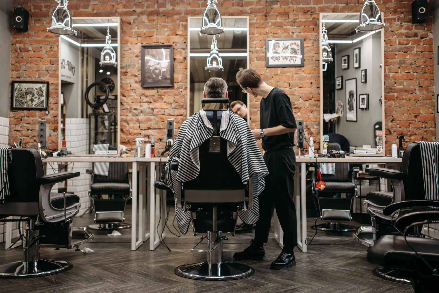Barbero atendiendo a un cliente en una barbería con paredes de ladrillo visto y sillones clásicos