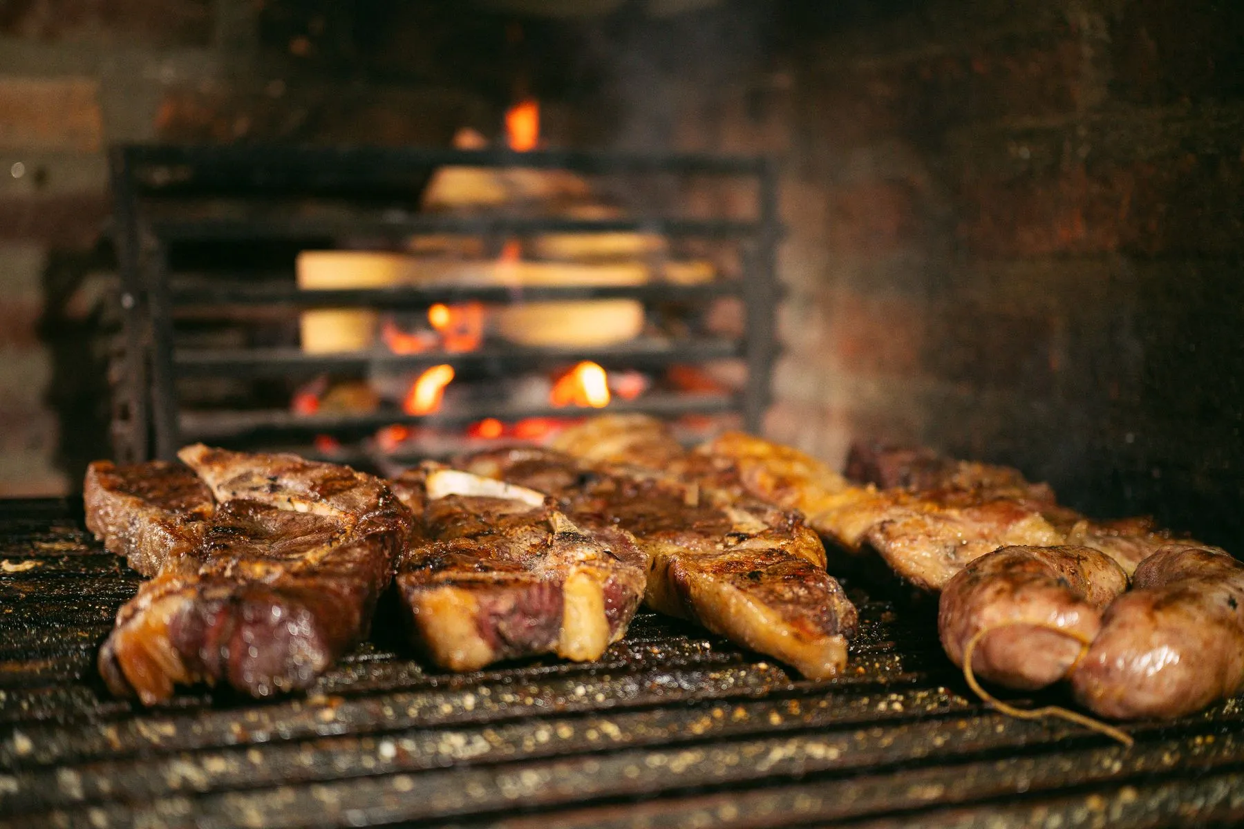 Cortes de carne asándose sobre la parrilla con el fuego de leña al fondo