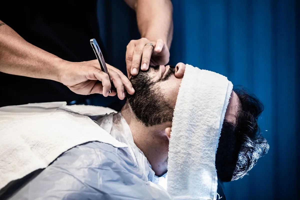 Cliente recostado con toalla caliente mientras el barbero le perfila la barba